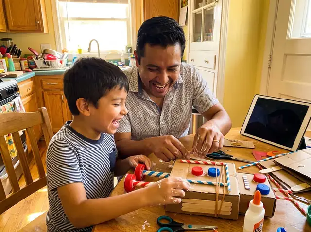 Pai e filho rindo enquanto montam um robô de papelão na mesa da cozinha